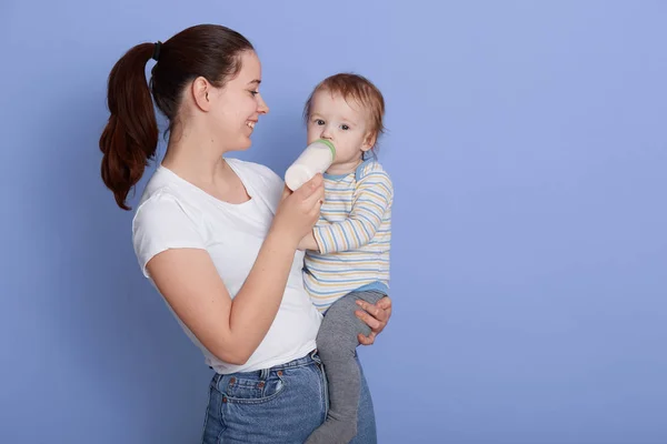 Retrato de una mujer caucásica sosteniendo a su bebé mientras se alimenta de biberón, posando aislado ove rblue fondo, señora mirando a su hijo en brazos, usando ropa casual, expresando felicidad . — Foto de Stock