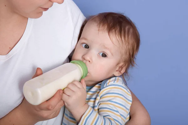 Hirizontal πλάνο του liitle γοητευτικό μωρό πόσιμο γάλα του μπουκαλιού, είναι στην αγκαλιά της μητέρας, αισθάνεται πείνα, κοιτάζοντας στην άκρη, φορώντας ριγέ πουκάμισο, βρεφική χαλάρωση και αίσθημα ευτυχίας κοντά στη μαμά της. — Φωτογραφία Αρχείου