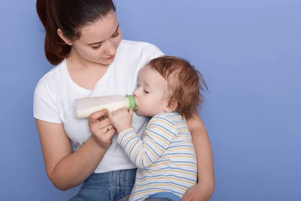 Picture of attarctive young mum feeding her little son from small bottle, mother holding kid in arms while posing isolated over blue studio background, baby dresses striped shirt. Childhood concept. — Stock Photo, Image