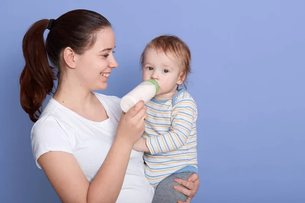 Close up portrait of younge brynette mother feeding her baby isolated over blue background, attractive female with ponytail, wearing white casual t shirt holding her little child and smiling. — Stock Photo, Image