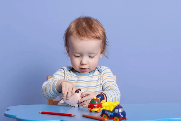 Retrato de cerca del pequeño chico caucásico. Lindo bebé sentado en el escritorio del niño y jugando con el juguete por sí mismo. Adorable niño siendo feliz y excitar, lleva camisa desnudada, aislado sobre fondo azul . —  Fotos de Stock