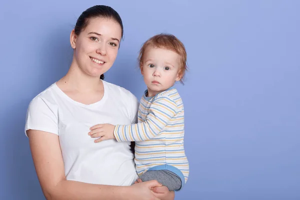 Studio shot of beautiful young mother standing and holding her baby boy in her arms in front of blue background, lady with dark hair and ponytail with cute infant, looking ar camera and smiling. — Stock Photo, Image