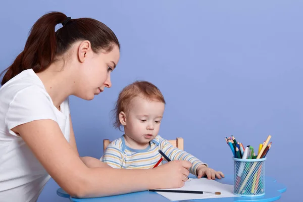 Horizontale Aufnahme von Kind und Mutter, die mit bunten Bleistiften zeichnen, während sie als Kind zu Hause sitzen, Mama, die Zeit mit ihrem Kind verbringt, mun und Kind wirken konzentriert, machen Geschenk für Vater. — Stockfoto