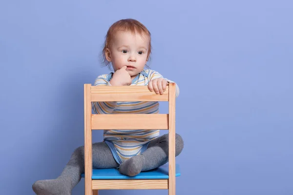 Horizontal shot of little boy sitting on stool, isolated on blue studio background, looks thoughtfuly aside, keeps finger near lip, infant wearing striped bodysuit and gray tights. Childhood concept. — Stock Photo, Image