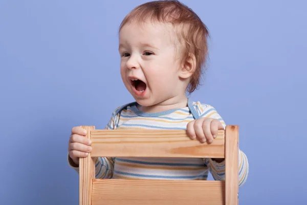 Tiro horizontal de menino feliz sentado na cadeira da criança, bebê adorável vestindo bodysuit, gritando algo em voz alta, bebê bonito posando isolado sobre o fundo do estúdio azul. Conceito de infância . — Fotografia de Stock