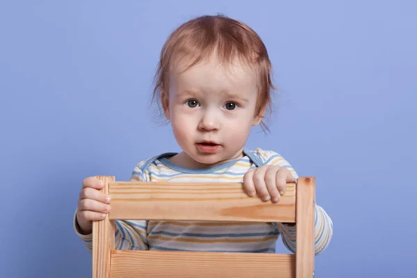 Tiro horizontal de criança liitle bonito sentado com expressão facial animado na cadeira de madeira, criança vestindo bodysuit, charmoso bebê posando isolado sobre fundo bluestudio. Conceito de infância . — Fotografia de Stock