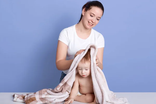 Horizontal shot of cute happy baby kid sitting wrapped in white and brown towel after bath, mum wiping and drying kid isolated over blue background, smiling female looking at her child woth gentle. — Stock Photo, Image