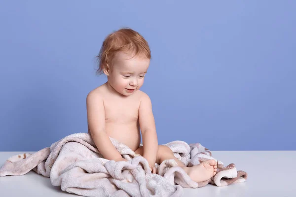 Οριζόντια λήψη του γοητευτικό αγοράκι κάθεται στην πετσέτα απομονώνονται σε μπλε φόντο, αξιολάτρευτο βρέφος μετά το μπάνιο, γυμνό cheeper παίζει και κοιτάζοντας γυμνό πόδι του με ενδιαφέρον. Έννοια της παιδικής ηλικίας. — Φωτογραφία Αρχείου