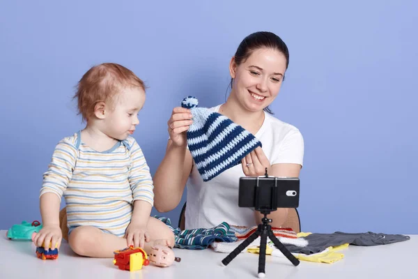 Close up portrait of contemporary young woman holding knitted cap while filming video for fashion and beauty channel against blue background, posing with her child isolated over blue background. — Stock Photo, Image
