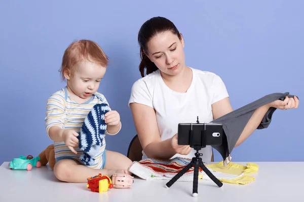 Image of fashion blogger shooting new video for her vlog tjgether with her little son. Beautiful woman showing clothes to camera and sitting isolated over blue studio background. Vlogging concept. — Stock Photo, Image