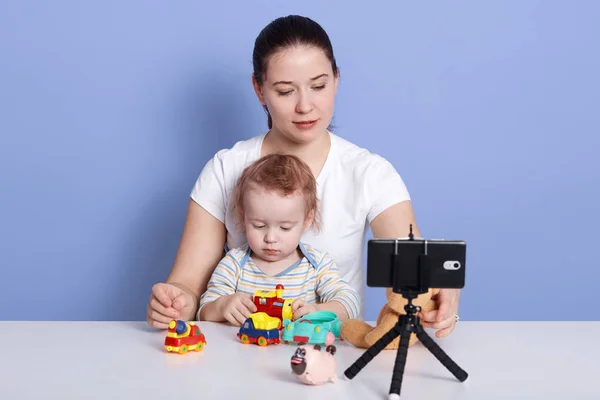 Woman vlogger looking using tripod and phone filming video about her little son and life. Girl sharing on internet social networks lifestyle contents, posing with cute infant in front of camera. — Stock Photo, Image