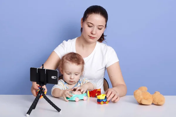 Mother and son playing with toys isolated over blue studio background, making content for her blog, infant being fond of car toy, looks concentrated and interested in playing. Blogging concept. — Stock Photo, Image