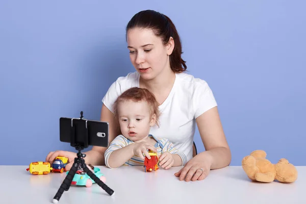 Close up portrait of baby boy and his mother playing together with toys, making video for vlog, posing isolated over blue studio background, attractive mom and child in front of tripoid with phone. — Stock Photo, Image