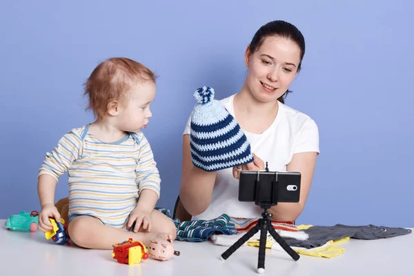 Horizontal shot of young woman holding knit cap while filming video for her channel against yblue background, sitting in front of camera with infant boy wears stripped bodysuit. Vlogging concept. — Stock Photo, Image
