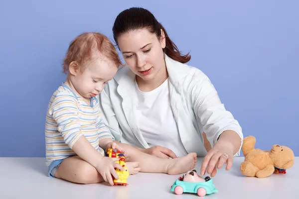Close up portrait of mother and baby boy having fun with toys, sitting isolated over blue background, infant wearing stripped bodysuit looks concentrated, mom and son with serious facial expression. — Stock Photo, Image