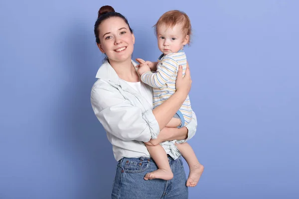 Indoor portrait of young mother holding her infant son in arms, child wearing stripped bodysuit, lady dresses white shirt, darkhaired girlwith her baby posing isolated over blue studio background. — Stock Photo, Image