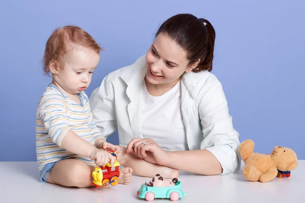 Horizontal shot of mother and child son being engaged in creativity in studio over blue background, mummy looking smiling at her charming child, cute kid playing with car toys. Motherhood concept. — Stock Photo, Image