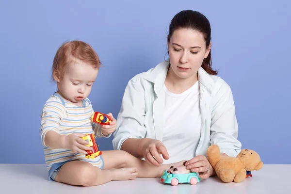 Horizontal shot of mom with kid son play together in room at home, infant wearing striped bodysuit, dark haired mother dresses casual clothing, family posing isolated over blue studio background. — Stock Photo, Image