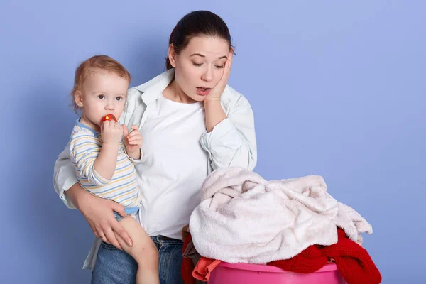 Plano horizontal de joven hermosa madre sosteniendo bebé niño en sus brazos mientras mira ropa sucia en el lavabo, necesidadesponer ropa de cama en la lavadora, mamá y bebé aislado sobre fondo azul . — Foto de Stock