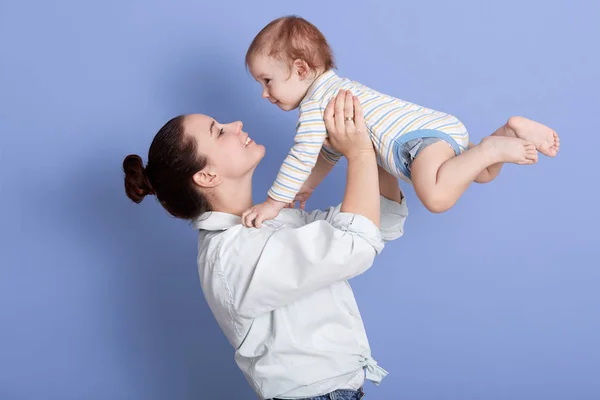 Indoor shot of mother playing with baby boy, happy family having fun at home, cheerful sweet kid in mom 's arms, mom and child isolated over blue background, mummy lifting toddler above her head . — Foto de Stock