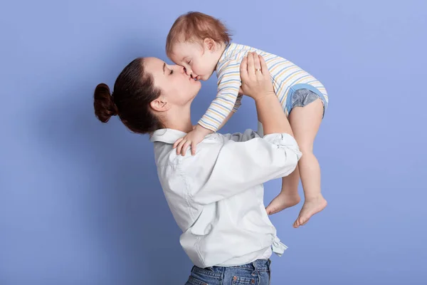 Horizontal shot of mom throws baby and kiss his, playing together and having fun, mother wearing white shirt and jeans, infant dresses stripped bodysuit, posing isolated over blue studio baground. — Stock Photo, Image