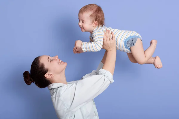 Portrait of happy mother and child together having fun isolated over blue background, attractive female with bun, dresses white shirt, infant wearing striped bodysuit. Mom and son, family concept. — Stock Photo, Image