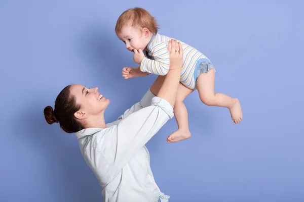 Side view of mid adult Caucasian mom lifting happy toddler son into air above head, attarctive mommy wearing white shirt, having dark hair with bun, happy child spending time with his beloved mom. — Stock Photo, Image