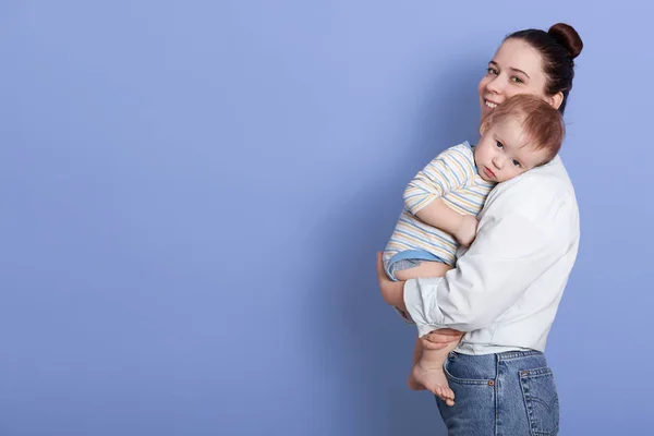 Retrato de cerca de la hermosa madre joven de pie y sosteniendo a su bebé en brazos, usando ropa casual, bebé acostado sobre los hombros de mamá. Copiar espacio para publicidad o texto promocional . — Foto de Stock
