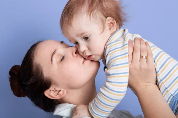 Horizontal shot of mom throws up baby and kissing, playing and having fun, brunette female with bun lifting infant dresses stripped bodysuit, isolated over blue studio background. Family concept. — Stock Photo, Image