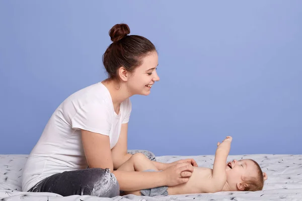 Foto horizontal de madre feliz posando con el bebé, mamá sentada en la cama cerca de su bebé, sonriendo bebé jugando con la amada madre, posando aislada sobre fondo azul del estudio. Familia, concepto de maternidad . — Foto de Stock