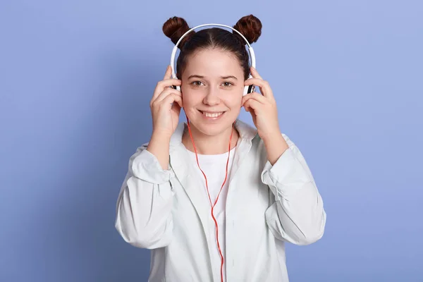 Bild der schönen Mädchen mit doppelter Frisur, die direkt in die Kamera schaut, während sie Musik über Kopfhörer hört, isoliert über blauer Studiowand, bezaubernde Frau in weißer Kleidung. — Stockfoto
