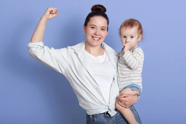 Retrato de madre sonriente con el bebé en brazos, mostrando sus bíceps a la cámara, muestra su fuerte, posando aislada sobre fondo azul del estudio, vistiendo ropa casual, hembra de pelo oscuro con moño . — Foto de Stock