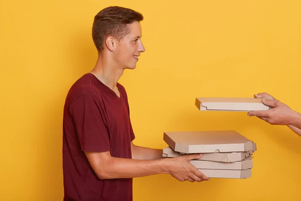 Profil de joyeux jeune garçon agréable tenant des boîtes de carton avec pizza, mains inconnues mettant une boîte de plus, souriant sincère, debout isolé sur fond jaune en studio. Concept de livraison — Photo