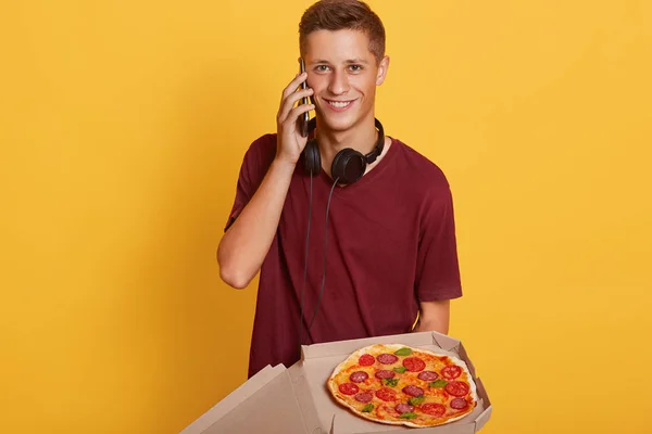 Plan horizontal d'un livreur sincère souriant parlant au téléphone, ayant un casque autour du cou, livrant une boîte à pizza, étant poli avec les clients, étant charismatique. Concept de livraison . — Photo