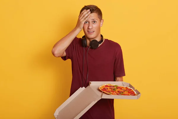 Imagen de un guapo y atractivo joven carismático parado aislado sobre un fondo amarillo, tocándose la frente con una mano, sosteniendo una caja de cartón con pizza, confundiéndose con el orden de los alimentos . — Foto de Stock