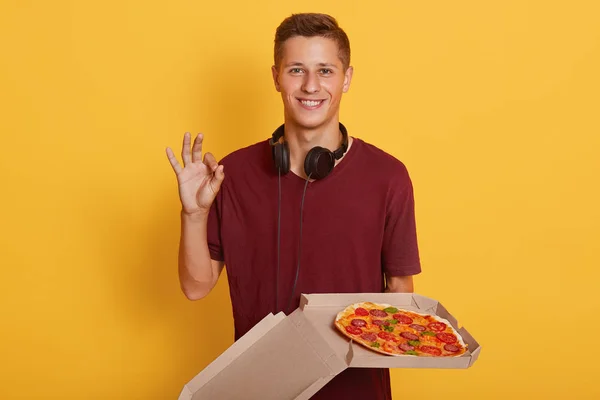 Photo de jeune garçon magnétique énergique tenant boîte ouverte avec pizza, ayant casque autour du cou, portant t-shirt rouge, debout isolé sur fond jaune en studio, montrant signe ok . — Photo