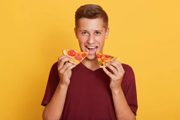 Indoor portret van grappige vrolijke jongeman geïsoleerd over gele achtergrond, twee stukken pizza in de mond, wijd open mond, het dragen van donker rood t-shirt. Voedselconcept. — Stockfoto