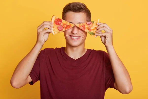 Beeld van vrolijke knappe jonge jongen met twee stukken pizza in beide handen, met een mooie glimlach, die zijn ogen bedekt met pizza, in een goed humeur, fastfood liefhebber. Concept mens en voedsel. — Stockfoto