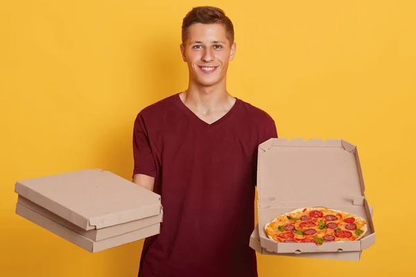 Retrato interior horizontal de un joven enérgico y atractivo mirando directamente a la cámara, con cajas con pizza en las manos, vistiendo una camiseta roja casual, sonriendo sinceramente. Concepto de entrega . — Foto de Stock