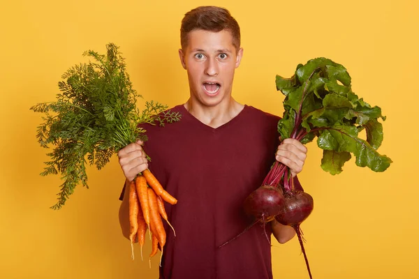 Elinde taze sebzeler olan yakışıklı bir adamın yatay görüntüsü, genç, çiğ yemek yiyen, sarı arka planda izole edilmiş, ağzı açık poz veren, hayret verici bir poz. Sağlıklı yeme kavramı.