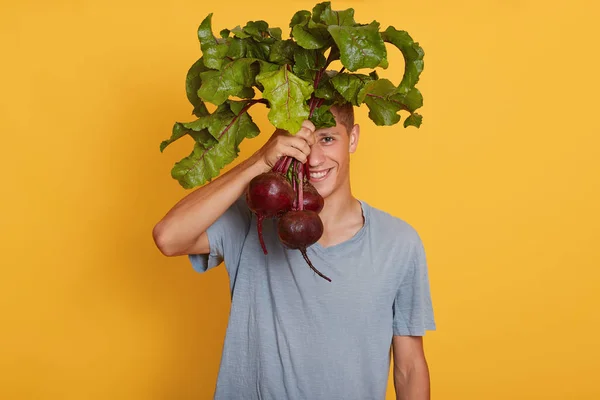 Plan horizontal de jeunes beaux mâles tenant des betteraves à la main, se cachant derrière des légumes frais, posant en tenue décontractée, couvrant son visage de betteraves. Aliments crus et concept de saine alimentation . — Photo