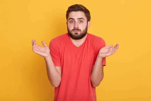 Pochybný emocionální pohledný mladík stojící osamoceně nad žlutým pozadím, zvedající ruce, gestikulující, s zmateným výrazem ve tváři, oblečený do červené ležérní trička. Emoční koncept. — Stock fotografie