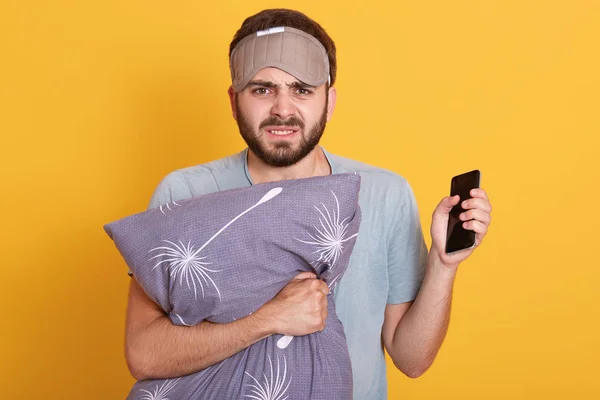 Närbild porträtt av arg irriterad ung man står isolerad över gul bakgrund i studio, med irriterad ansiktsuttryck, hålla pilow och smartphone. Vakna tidigt på morgonen koncept. — Stockfoto