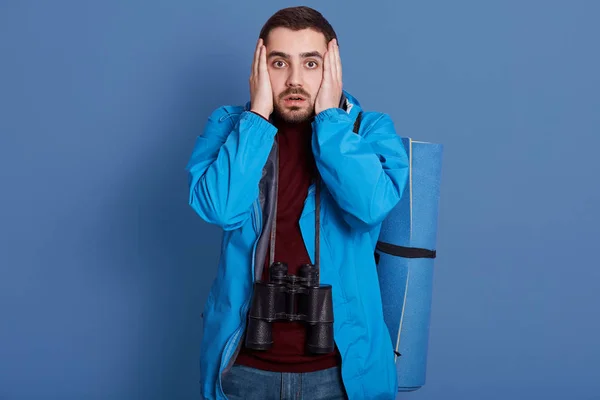 Horizontální vnitřní portrét emočního magnetického vousatého muže stojícího izolovaně nad přízračným modrým pozadím ve studiu, dotýkajícího se rukama svého obličeje, s dalekohledem kolem sebe a podložkou na spaní. — Stock fotografie