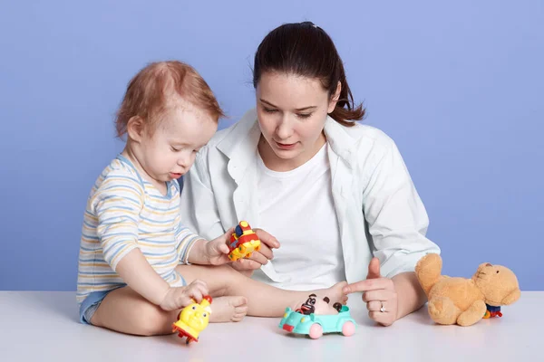 Close up portrait of young mother with her baby playing with toys, looking concentarted, female wearing casual clothing, infant dresse striped bodysuit, isolated over blue studio background. — Stock Photo, Image