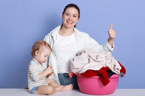 Indoor shot of mother with son sitting near laundry basin, look happy, young attractive female showing thumb up, being ready to work about house. Family, adorable kid, love and happiness concept. — Stock Photo, Image