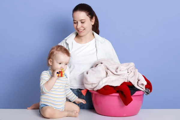 Indoor shot of charming infant boy and his mother sitting near basin full of dirty clothes, little child play weith toy, lady being ready to work, posing isolated over blue studio background. — Stock Photo, Image