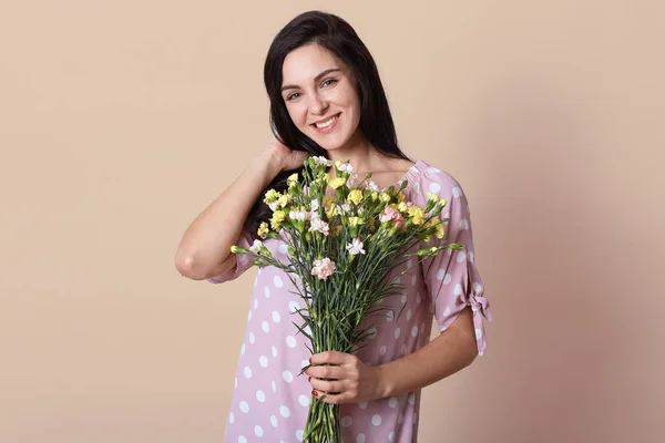 Horizontal shot of young woman in elegant rosy dress, posing isolated over beige background with bouquet in hands. Lady with spring flavor, looking smiling directly at camera, keeps hand on neck.