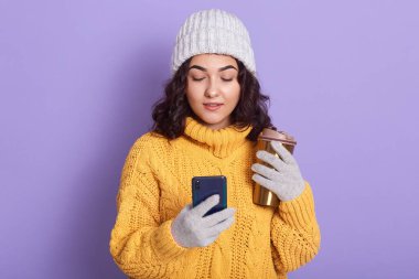 Gri şapkalı, eldivenli, elinde akıllı telefon ve elinde sıcak içecekle ekranda dikkatle duran güzel ve sevimli bir kadın resmi. Teknoloji konsepti.