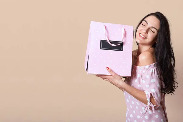 Image of beautiful tender peaceful female smiling sincerely, closing eyes, holding light pink package in both hands, being fond of shopping, being in high spirits. Copyspace for advertisement. — 스톡 사진